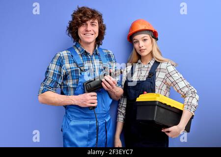 Two architects in uniform coveralls stand with tools instruments posing isolated on blue, young man hold drill in hands while his colleague woman hold Stock Photo
