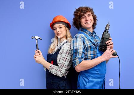 constructors builders stand back to back to each other holding household tools , team of positive builders man and woman in uniform and hardhat posing Stock Photo