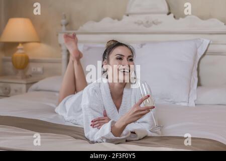 Happy female in white bathrobe smiling and drinking champagne while lying on soft bed in hotel room Stock Photo