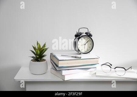 A stack of books, an artificial succulent plant in a ceramic pot, glasses, a metal basket with notebooks, a pen are on a white table surface. Concept Stock Photo