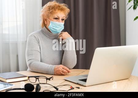 Elderly woman in medical mask works on laptop at home. Communication with friends and family remotely for old people. Insulation. Quarantine. Stock Photo