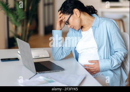 Exhausted sad pregnant mixed race adult woman sitting at her desk feels a headache, is stressed, holds her head with her hand, closing eyes, tired of sedentary work with laptop, need a break and rest Stock Photo