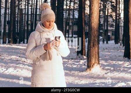 Middle-aged woman in warm white jacket and hat walks in winter park during sunset.Looks into the phone,reads messages,news,looks for new walking route Stock Photo