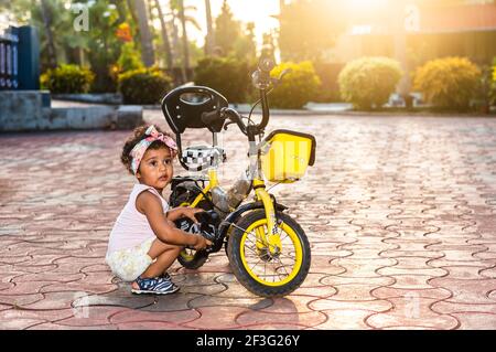 GOA, INDIA - May 12, 2020: Playful Pretty Indian girl child/infant/toddler wearing a hair band, playing with a cycle/tricycle. Kid giving joyful expre Stock Photo