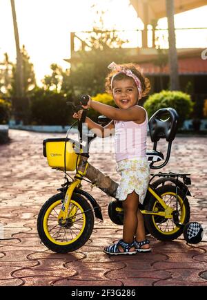 GOA, INDIA - May 12, 2020: Playful Pretty Indian girl child/infant/toddler wearing a hair band, playing with a cycle/tricycle. Kid giving joyful expre Stock Photo