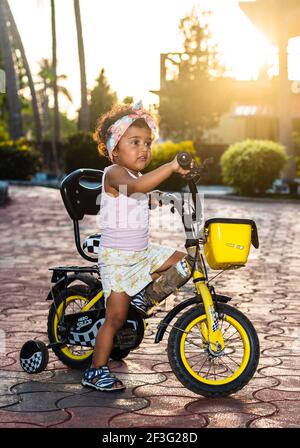 GOA, INDIA - May 12, 2020: Playful Pretty Indian girl child/infant/toddler wearing a hair band, playing with a cycle/tricycle. Kid giving joyful expre Stock Photo