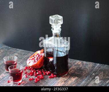 Pomegranate liqueur in a glass decanter and two glasses stands on a wooden table against a black wall, next to it is ripe broken fruit and scattered s Stock Photo