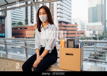 Stressed and worried young Asian woman with box of items sitting alone after being laid off from job due to covid-19 pandemic Stock Photo
