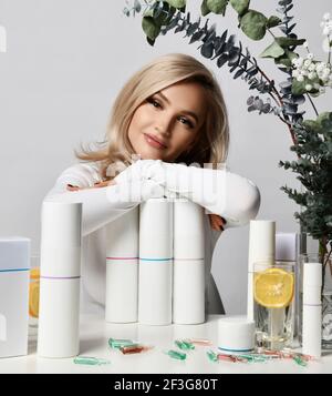 Healthy wealthy blonde woman in white stylish pullover sits at table leaning on set of jars and tubes of cosmetic means Stock Photo