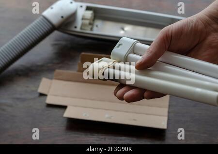 Adult Man's hand holds a burned-out and a new compact fluorescent bulb. Table lamp and cardboard packaging in the background. Replacing the burned-out Stock Photo