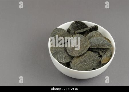 Black chips in a white ceramic bowl on a gray background. Salty chips with spices and chili peppers. Ready-to-eat snacks. Stock Photo