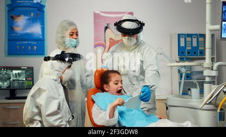 Happy little girl in ppe suit looking in the mirror after dental intervention sitting in new normal stomatological office. Dentist in coverall showing the result after hygienic cleaning examination Stock Photo