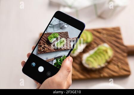 Hands take pictures on smartphone of two beautiful healthy sour cream and avocado sandwiches lying on board on the table. Social media and food Stock Photo