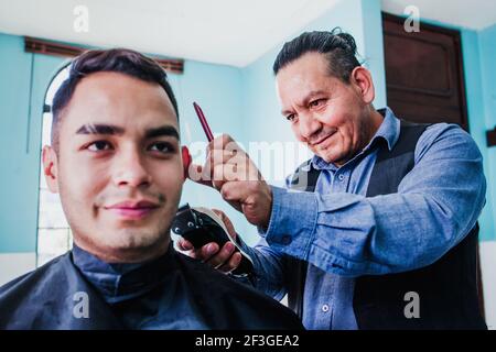 latin man stylist cutting hair to a client in a barber shop in Mexico Stock Photo