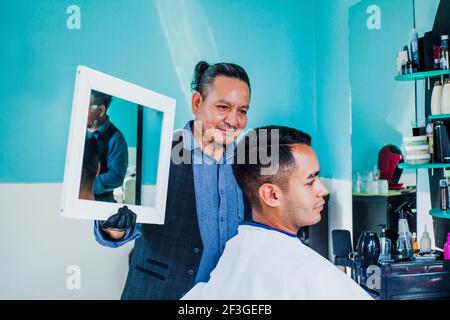 latin man stylist cutting hair to a client and holding a mirror in a barber shop in Mexico Stock Photo