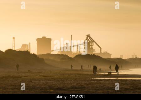 Redcar, Cleveland, UK, 16th March 2021. People walking and playing football on the beach at Redcar, Cleveland, UK, as the Sun sets behind the Teesside Steelworks. The Redcar site once contained the largest blast furnace in Europe. Due to the Chinese government flooding the world market with cheap subsidised steel, British steel became uneconomic. The Teesside site closed in 2015. Credit: Paul Thompson/Alamy Live News. Stock Photo