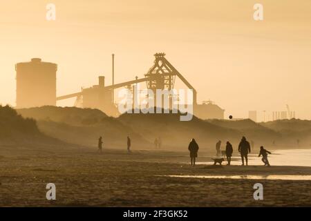 Redcar, Cleveland, UK, 16th March 2021. People walking and playing football on the beach at Redcar, Cleveland, UK, as the Sun sets behind the Teesside Steelworks. The Redcar site once contained the largest blast furnace in Europe. Due to the Chinese government flooding the world market with cheap subsidised steel, British steel became uneconomic. The Teesside site closed in 2015. Credit: Paul Thompson/Alamy Live News. Stock Photo