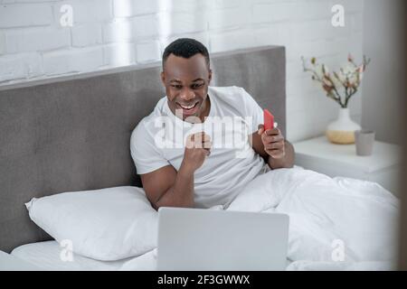 A young man in white tshirt lying in bed and working remotely Stock Photo
