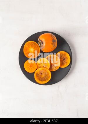 Ripe orange juicy persimmon slices and whole fruit on black plate on white textured background. Top view. copy space.