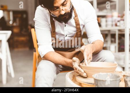 A young male potter is engaged in craft in his workshop on a potter's wheel and makes a clay product Stock Photo