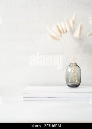 Still life of white dry lagurus flower in gray glass vase on stack of white magazines on light background. Front view. Copy space Stock Photo