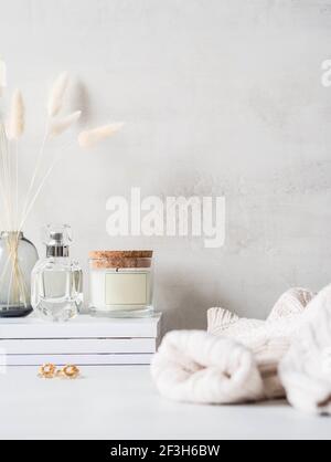 Modern feminine still life of sweater, white dry lagurus flower in glass vase and various items on stack of white magazines on light background. Front Stock Photo