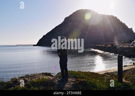 FRESNO, UNITED STATES - Mar 04, 2021: A photo of a male from behind standing on a hill looking outat the water and Morro Rock during sunset Stock Photo
