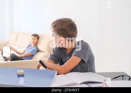 Boy doing homework with books notebooks and other boy using digital tablet for online lessons. Brothers at home talking about homework tasks Stock Photo