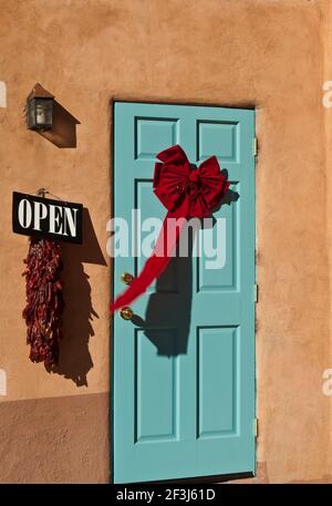 Close-up view of turquoise door with red ribon Christmas decoration in an adobe wall with open sign and chilli ristra. Stock Photo
