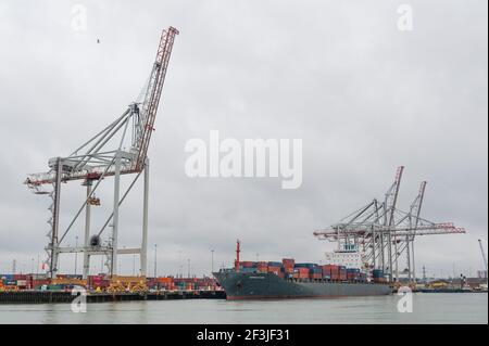 Southampton, UK, 04 Mar, 2021. Overcast skies provide a gloomy backdrop to part of Southampton dock yard where a cargo ship loaded  with metal shipping containers is berthed next to giant dockyard cranes. Southampton docks has recently been given free port status. Stock Photo