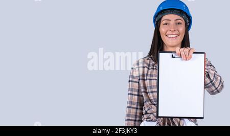 Banner, long format. Cheerful construction engineer, caucasian woman with hard hat holding clipboard with blank white sheet for ads, grey background. Stock Photo