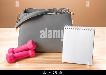 Women leather grey handbag, notepad and small pink dumbbells. women's bag and Notepad for writing and fitness dumbbells. concept schedule training, pl Stock Photo