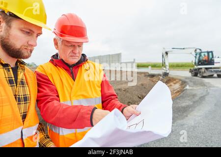 Architect and construction worker with floor plan when building a house on a construction site with excavator Stock Photo