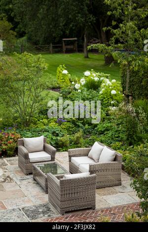 York stone paved evening seating area and herring-bone red brick detail with cushioned, cane furniture adjacent to bedding and the lawn at the rear of Stock Photo