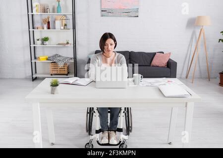 Handicapped freelancer using laptop near documents with charts and cup on table Stock Photo