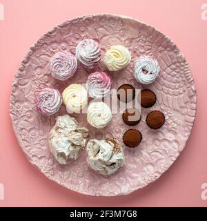Pink ceramic plate with chocolate truffles, meringues, marshmallow cupcakes on pink background. Close up, top view. Stock Photo