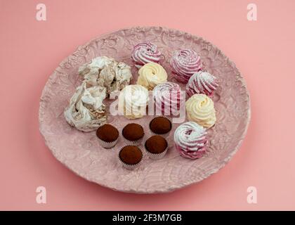 Pink ceramic plate with chocolate truffles, meringues, marshmallow cupcakes on pink background. Close up. Stock Photo