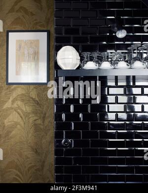 Brick tiled wall with glassware and crockery on shelf next to gold metallic patterned wallpaper in French residential home. Stock Photo