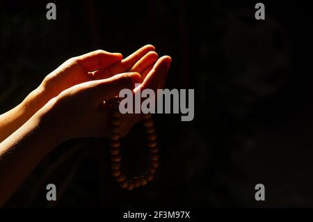 Hands of a child praying with a prayer beads on a dark background and backlight Stock Photo