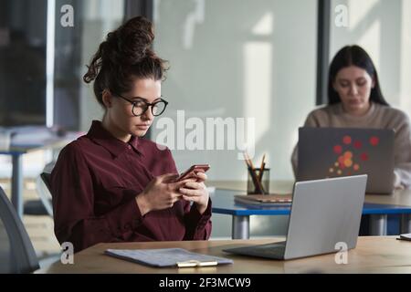 Young businesswoman in eyelasses sitting at her workplace with laptop and using mobile phone Stock Photo