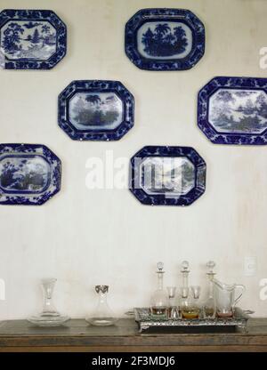 Collection of china plates mounted on wall with decanters and glasses on side table in residential house, Brazil. Stock Photo