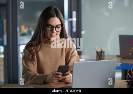 Young woman in eyeglasses reading a message on mobile phone at her workplace at office Stock Photo
