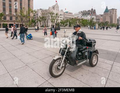 Shanghai, China - May 4, 2010: Closeup of male senior on motorized tricycle riding on boardwalk along Bund. Iconic historic buildings in back. Clothin Stock Photo