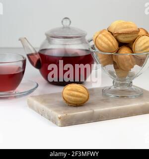 Homemade nut-shaped cookies stuffed with condensed milk in a glass vase on a marble cutting board. Red Sudanese rose herbal tea in a cup and teapot. Stock Photo