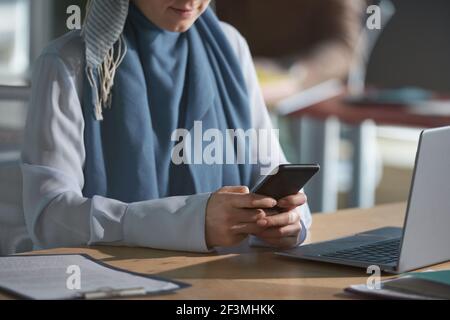 Close-up of young woman in hijab sitting at her workplace with laptop and using mobile phone at office Stock Photo