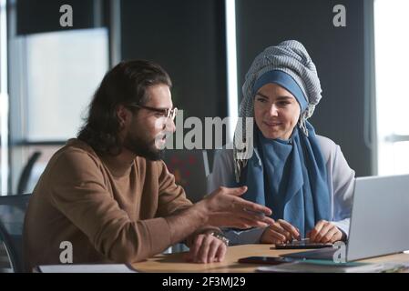 Businessman talking to his colleague in hijab they using laptop sitting at the table at office Stock Photo