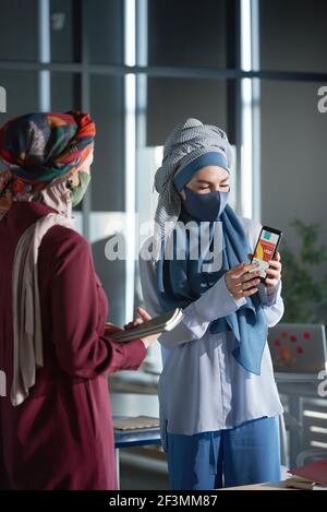 Two muslim businesswomen in hijabs using application on mobile phone during work at office Stock Photo