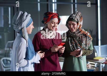 Three muslim businesswomen in hijabs using mobile phones and discussing mobile apps while standing at office Stock Photo