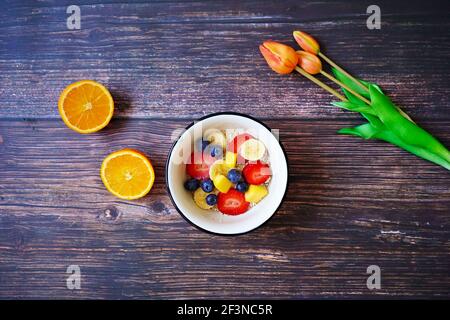 A cereal bowl with fresh fruits (strawberry, banana, blueberry, mango) and an orange on a wooden breakfast table. Orange tulips for decoration. Stock Photo