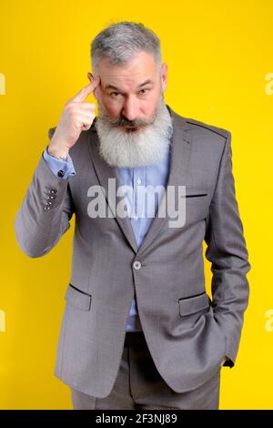 Studio portrait mature businessman dressed in gray suit pointing to head with forefinger, great idea or thought concept, good memory, yellow backgroun Stock Photo